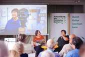 Franziska Gerstenberg liest vor Publikum aus einem Buchmagazin. Hinter ihr wird auf einer Leinwand ein Foto projiziert. Daneben ist das Logo der Kulturstiftung auf einem aufgestellten Banner zu sehen.