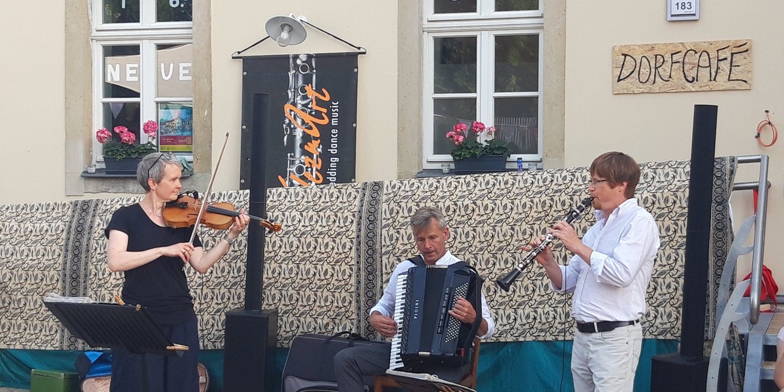 Drei Musizierende spielen gemeinsam vor einem Haus mit der Aufschrift "Dorfcafé".